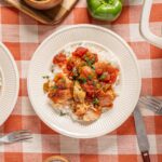 Sautéed chicken and vegetables served over rice on a white plate with fresh chopped herbs, set on a red and white checkered tablecloth with silverware and a green bell pepper in the background.