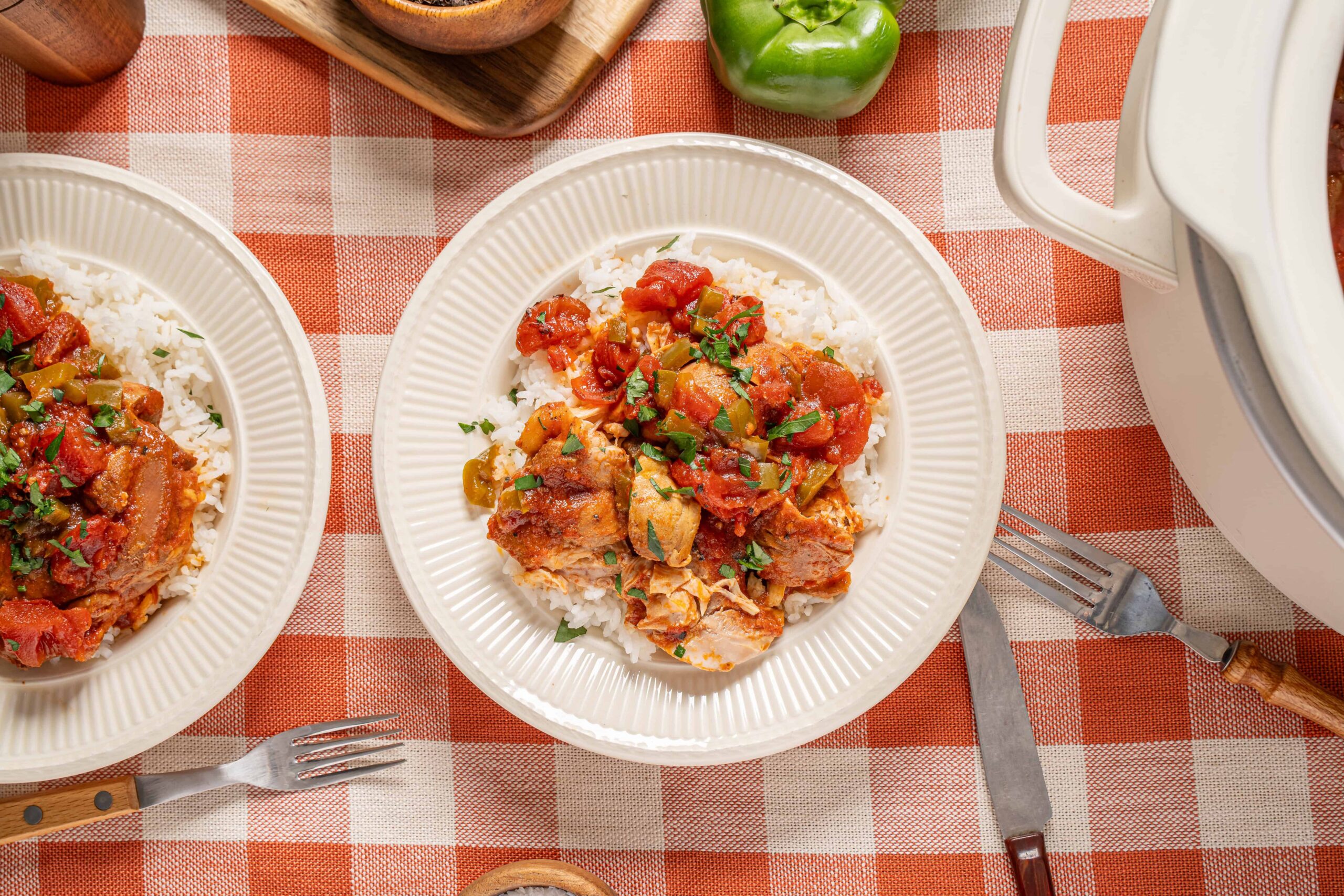Sautéed chicken and vegetables served over rice on a white plate with fresh chopped herbs, set on a red and white checkered tablecloth with silverware and a green bell pepper in the background.