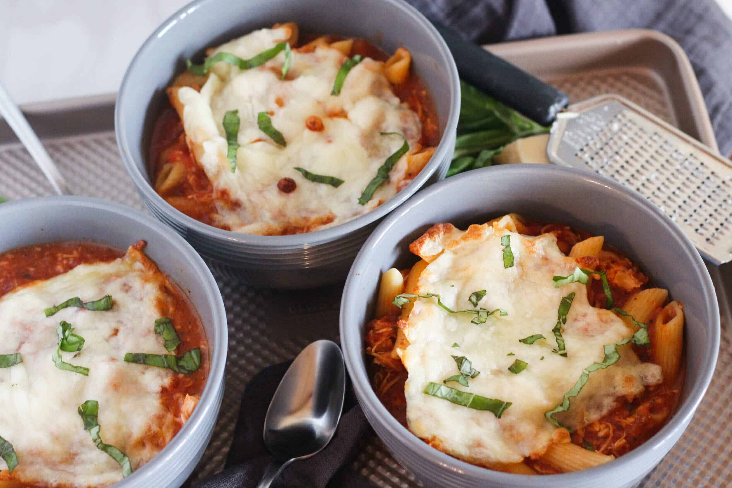 Creamy baked pasta casserole topped with melted cheese and fresh basil in gray bowls, served with a grater and fresh greens on a tray.