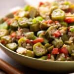 Sautéed okra with diced tomatoes, onions, and green onions in a bowl on a wooden surface, showcasing a healthy and flavorful vegetable dish.