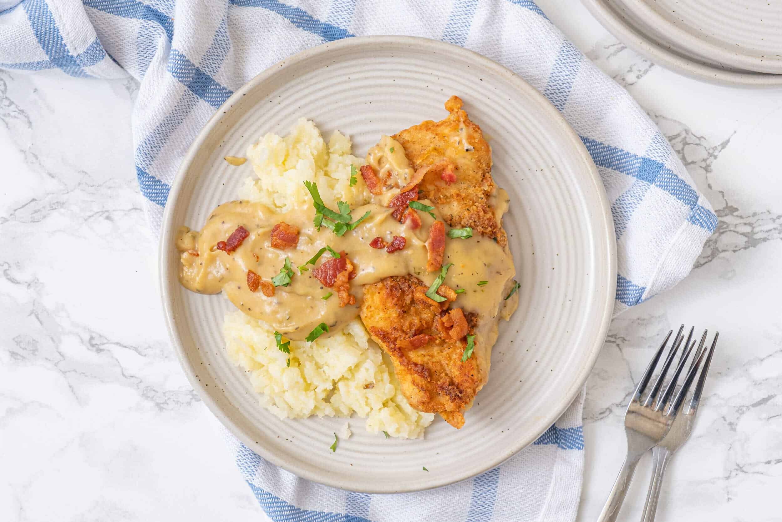Creamy mashed potatoes with chicken fried steak topped with gravy and bacon bits, served on a beige plate. Perfect comfort food dinner with crispy chicken and smooth mashed potatoes.