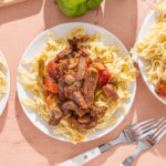 Juicy beef stew served over homemade pasta on white plates, with fresh green bell peppers in the background, perfect for comforting family meals or Sunday dinner.