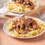 Savory beef and vegetable pasta served on a white plate, topped with tender beef strips, tomatoes, and peppers, alongside fettuccine noodles, perfect for a hearty Italian-inspired meal.