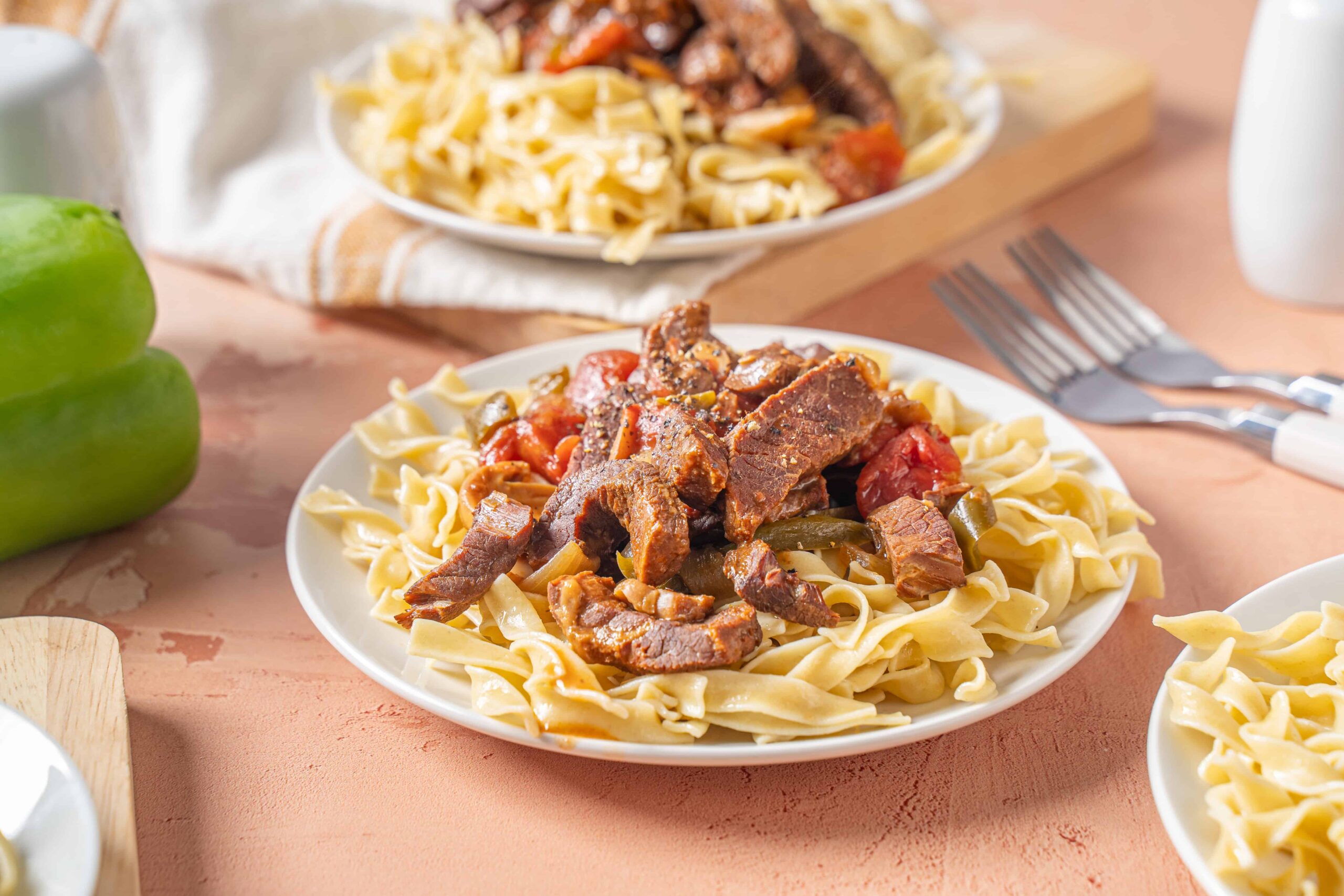 Savory beef and vegetable pasta served on a white plate, topped with tender beef strips, tomatoes, and peppers, alongside fettuccine noodles, perfect for a hearty Italian-inspired meal.