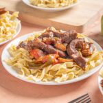 Sautéed beef and vegetable pasta on white plate with green bell pepper in background, close-up shot of hearty homemade beef pasta dish.