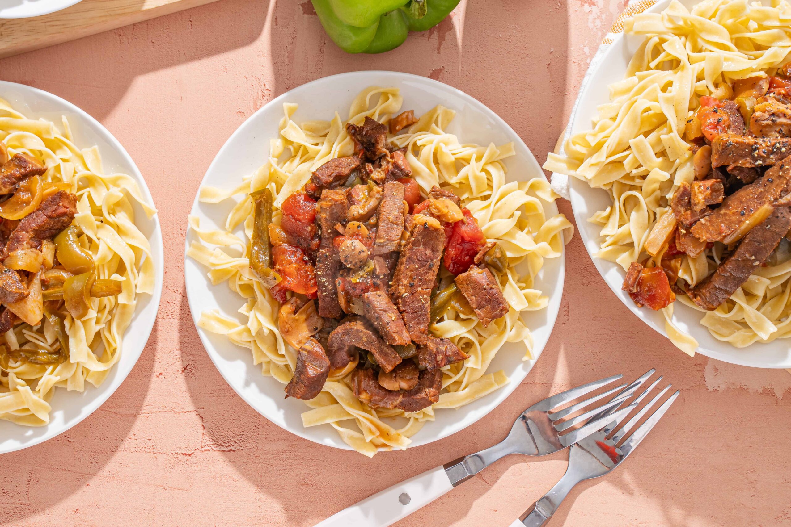 Juicy beef stew served over homemade pasta on white plates, with fresh green bell peppers in the background, perfect for comforting family meals or Sunday dinner.