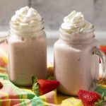 Creamy strawberry milkshake in mason jars topped with whipped cream, surrounded by fresh strawberries on a colorful cloth. Perfect for summer or snack cravings.