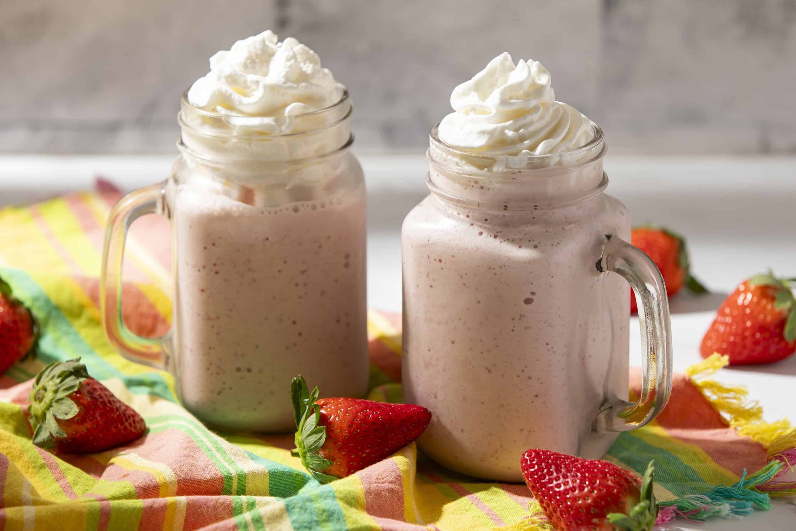 Creamy strawberry milkshake in mason jars topped with whipped cream, surrounded by fresh strawberries on a colorful cloth. Perfect for summer or snack cravings.