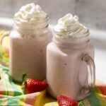 Creamy strawberry smoothies topped with whipped cream served in glass jar mugs with fresh strawberries around, on colorful cloth background for a delicious summer beverage.