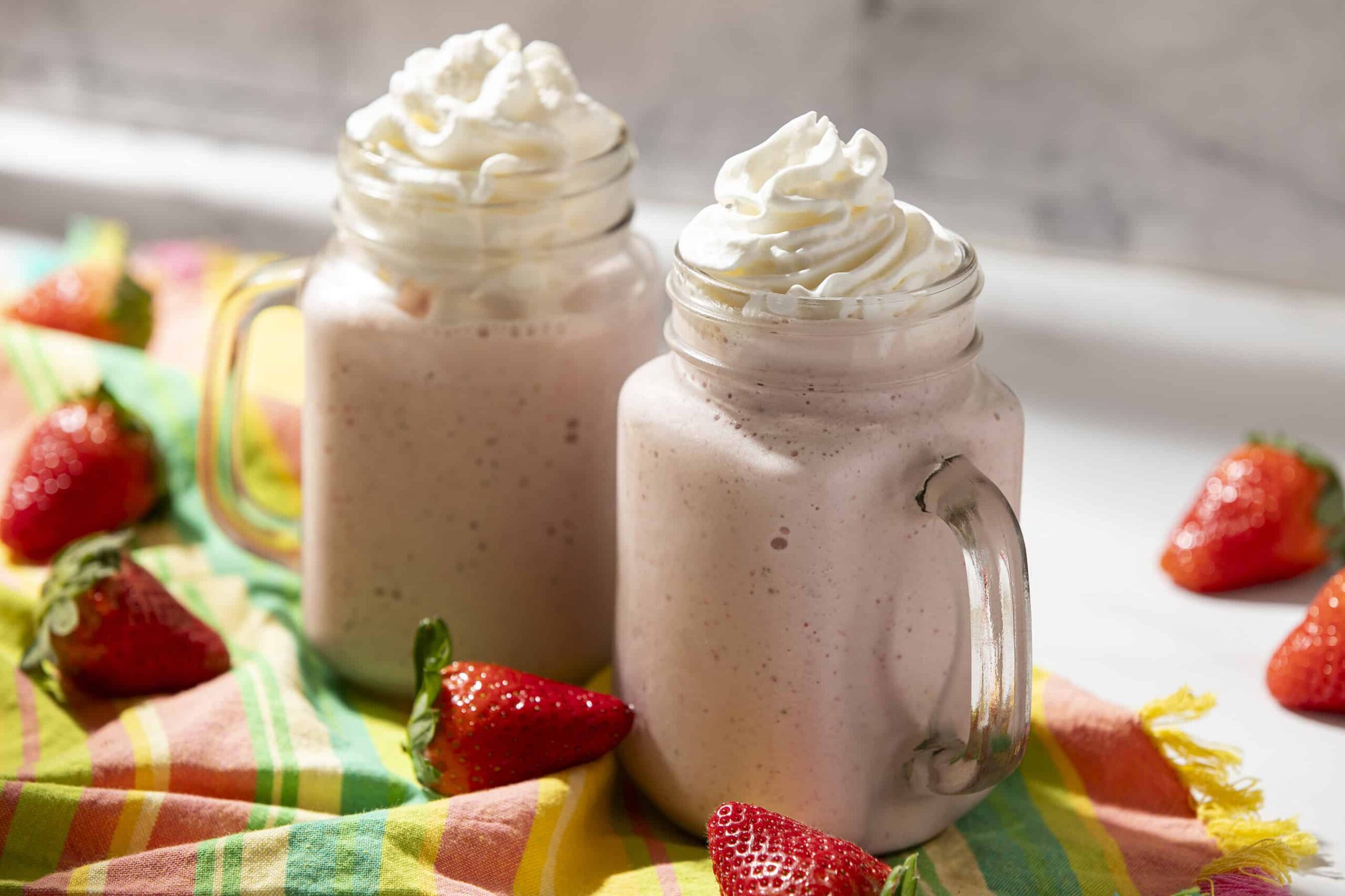 Creamy strawberry smoothies topped with whipped cream served in glass jar mugs with fresh strawberries around, on colorful cloth background for a delicious summer beverage.