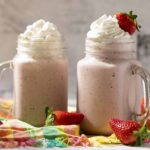 Creamy strawberry milkshake topped with whipped cream and fresh strawberries in mason jars, served on a colorful cloth with extra strawberries for a delicious summer beverage.