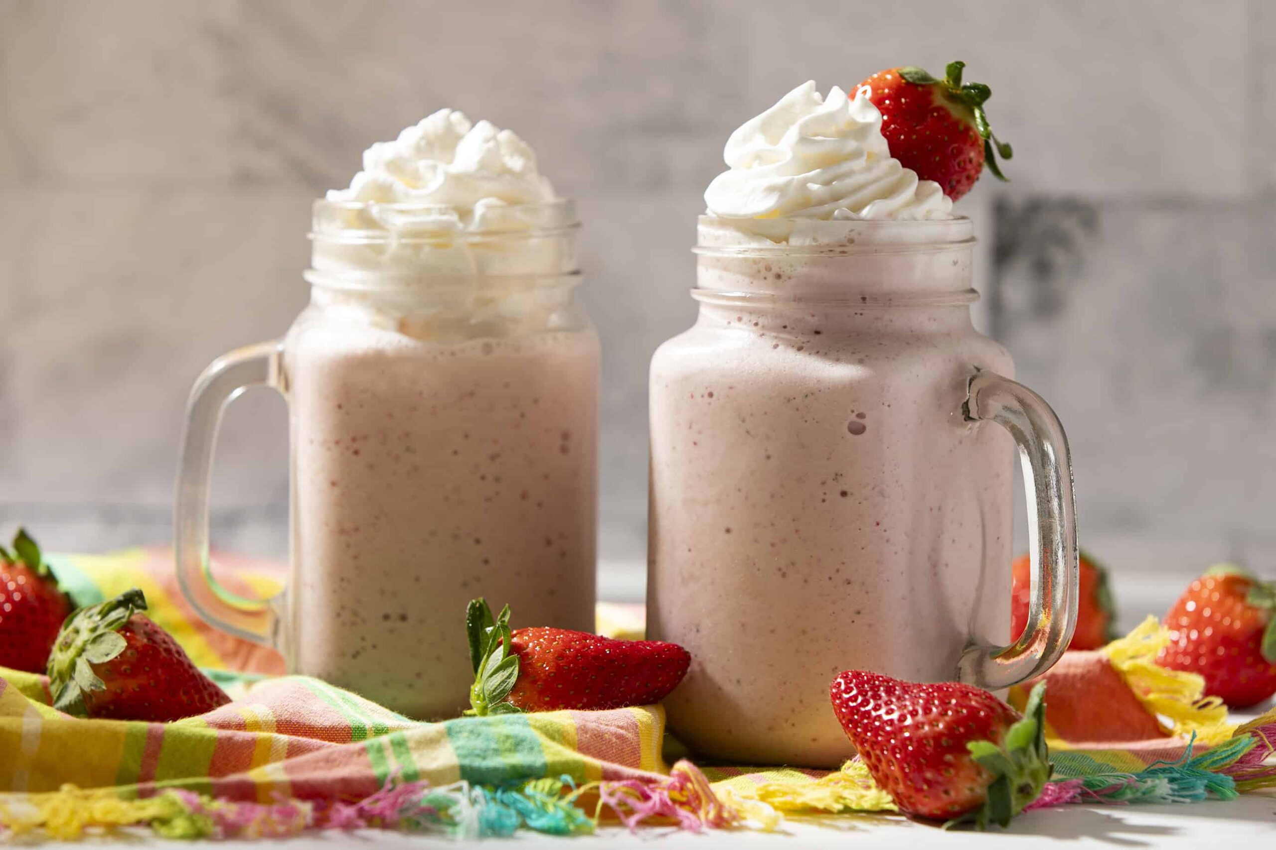 Creamy strawberry milkshake topped with whipped cream and fresh strawberries in mason jars, served on a colorful cloth with extra strawberries for a delicious summer beverage.