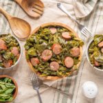 Sautéed collard greens with sausage served in white bowls, fresh green salad on side, rustic wooden utensils, and a linen tablecloth for a hearty, nutritious meal.