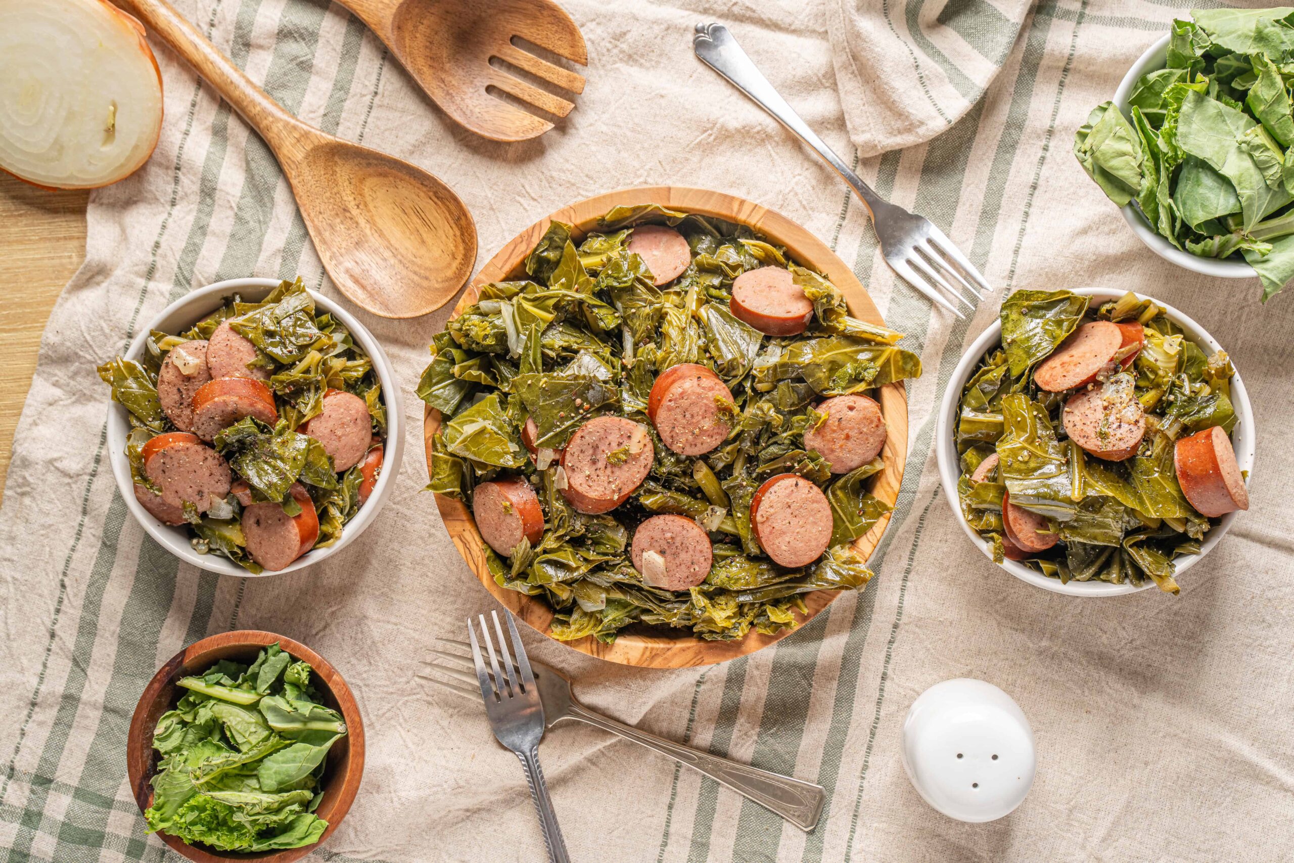 Sautéed collard greens with sausage served in white bowls, fresh green salad on side, rustic wooden utensils, and a linen tablecloth for a hearty, nutritious meal.