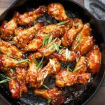 Crispy buffalo chicken wings in black cast iron skillet garnished with green onions and sesame seeds on wooden table.