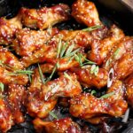 Sweet and spicy chicken wings glazed with a sticky sauce, garnished with sesame seeds and green onions, served in a black skillet, perfect for appetizers or party snacks.