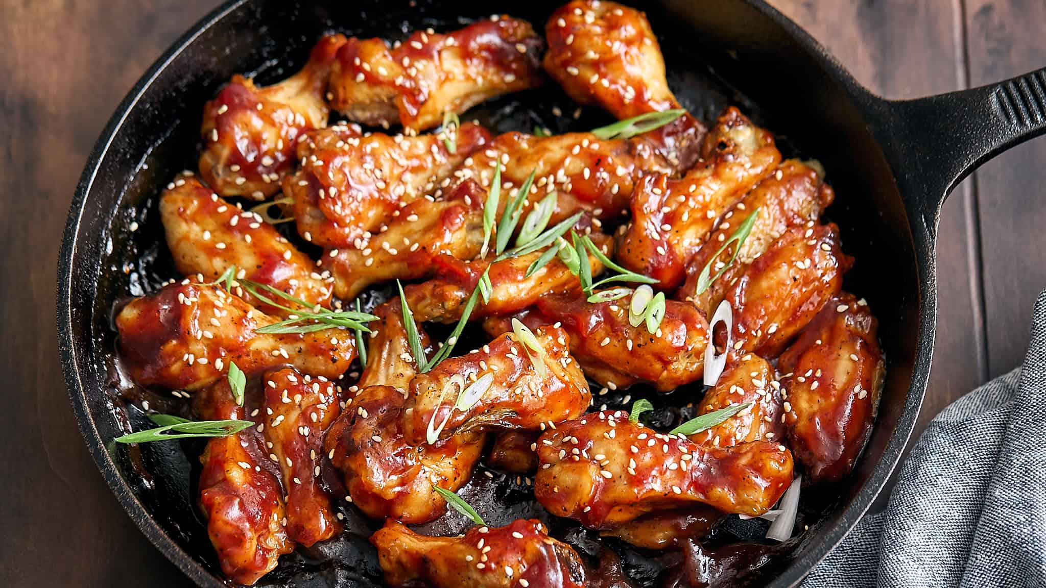 Sweet and spicy chicken wings glazed with a sticky sauce, garnished with sesame seeds and green onions, served in a black skillet, perfect for appetizers or party snacks.