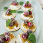 Miniature baked brie bites topped with cranberry sauce, fresh mint, and a dusting of powdered sugar on a light green platter. Perfect for holiday appetizers and entertaining.