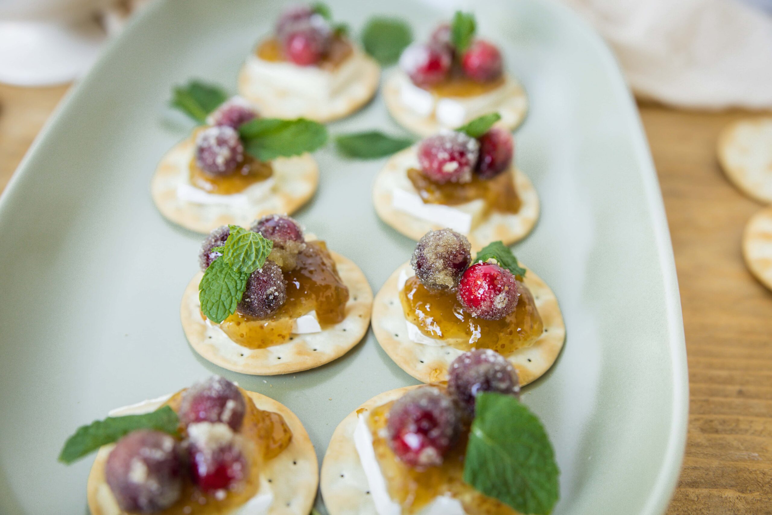 Miniature baked brie bites topped with cranberry sauce, fresh mint, and a dusting of powdered sugar on a light green platter. Perfect for holiday appetizers and entertaining.