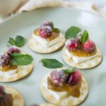 Mini crackers topped with cream cheese, preserved fruit, and fresh berries, garnished with mint leaves on a light green plate for a delicate appetizer or snack photo.