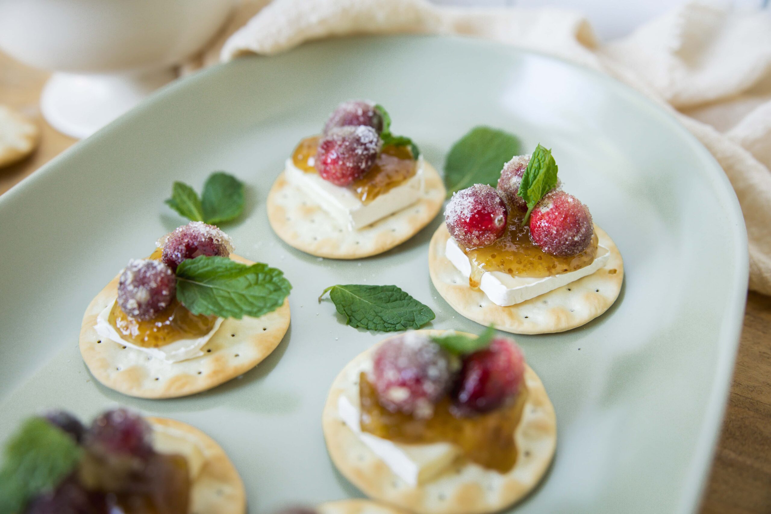 Mini crackers topped with cream cheese, preserved fruit, and fresh berries, garnished with mint leaves on a light green plate for a delicate appetizer or snack photo.