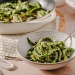 Fresh green bean salad with garlic and sesame seeds served in a white patterned bowl, with a larger dish of similar salad in the background, on a beige and wooden surface.