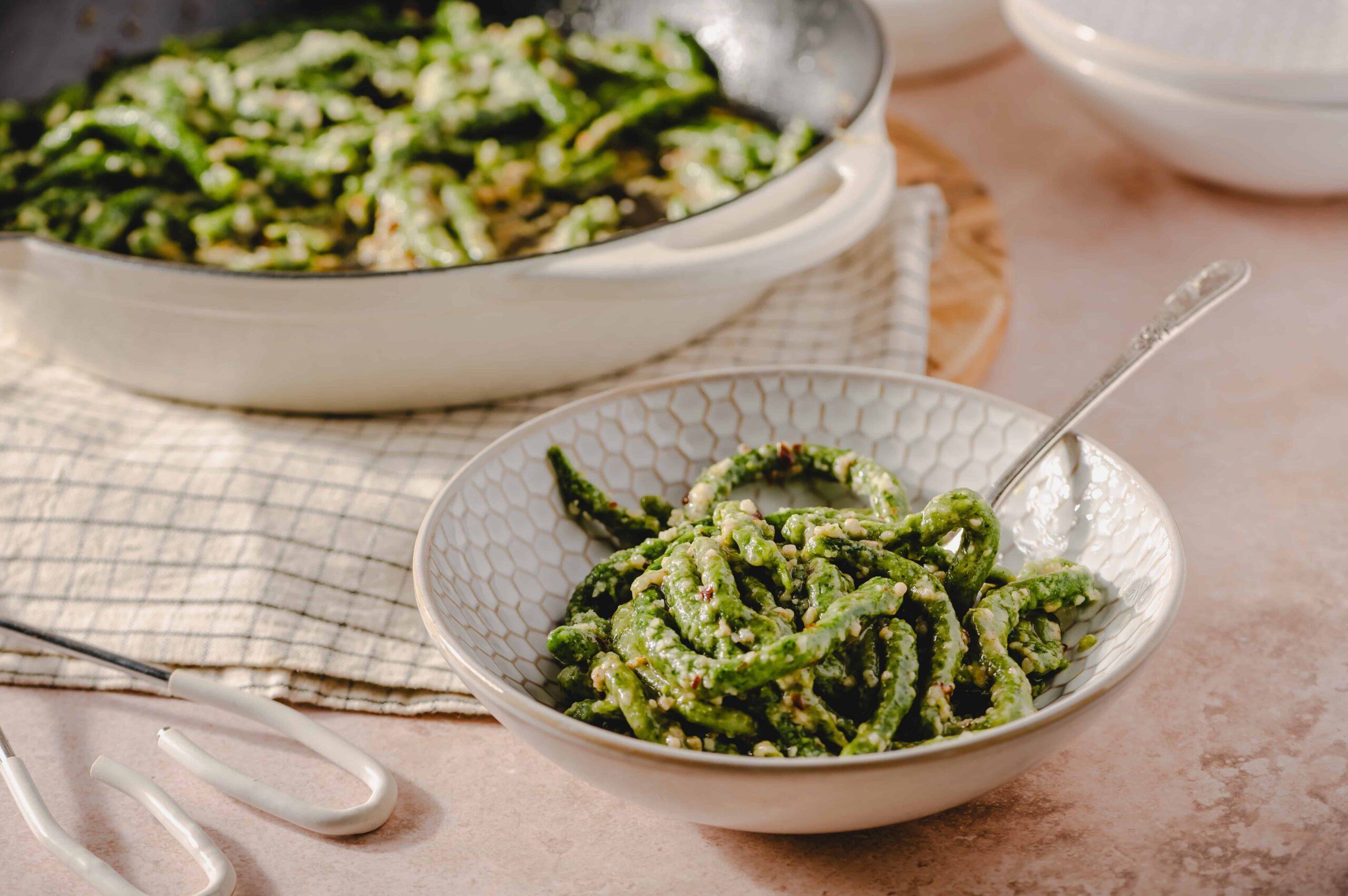Fresh green bean salad with garlic and sesame seeds served in a white patterned bowl, with a larger dish of similar salad in the background, on a beige and wooden surface.
