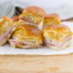 Buttery ham and cheese croissant sandwich squares with melted cheese and poppy seeds on top, served on parchment paper and wooden surface.