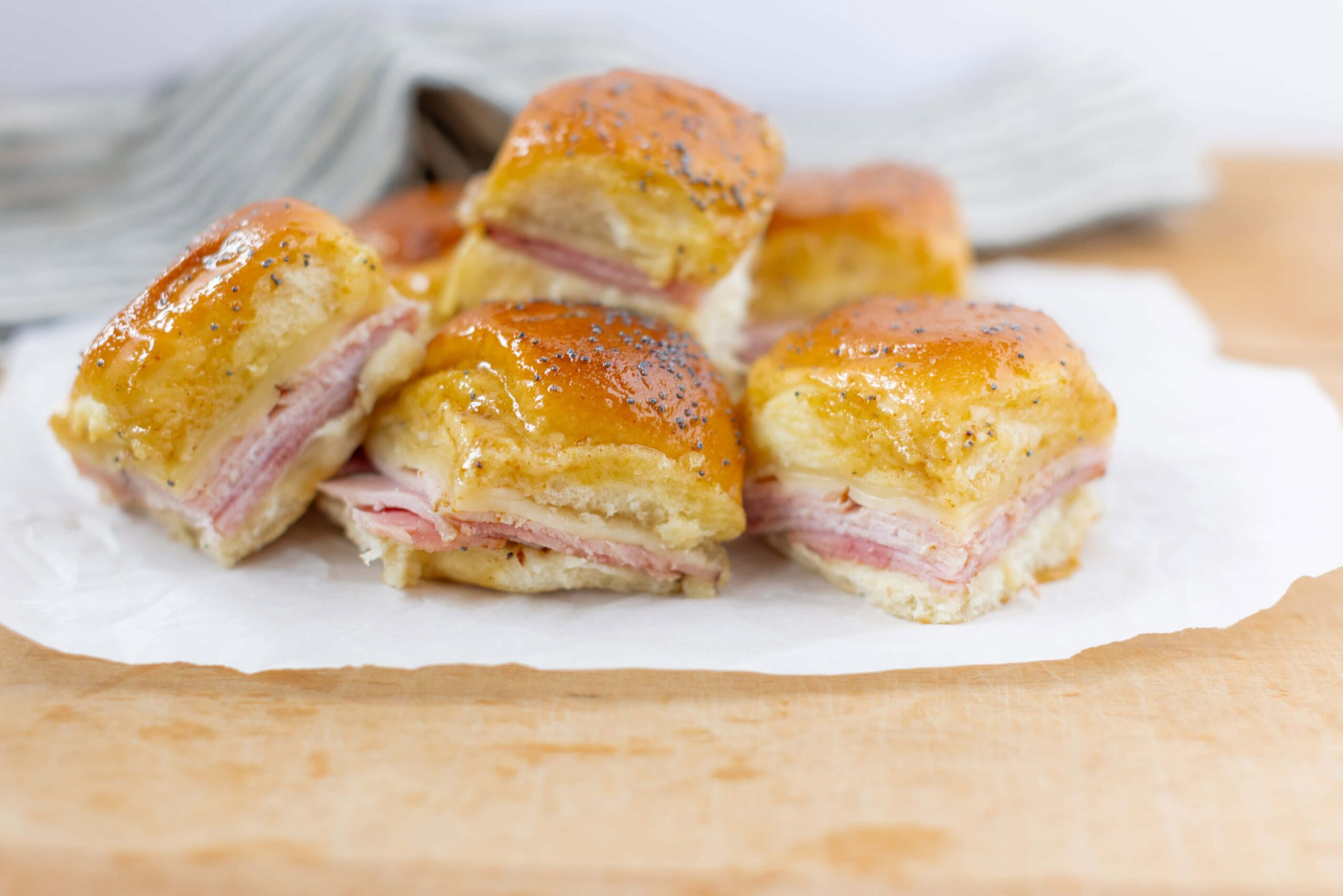 Buttery ham and cheese croissant sandwich squares with melted cheese and poppy seeds on top, served on parchment paper and wooden surface.