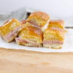 Buttered ham and cheese biscuits with a golden, flaky top, sprinkled with poppy seeds, served on parchment paper. Perfect for breakfast or snack, showcasing a savory pastry with ham and melted cheese filling.