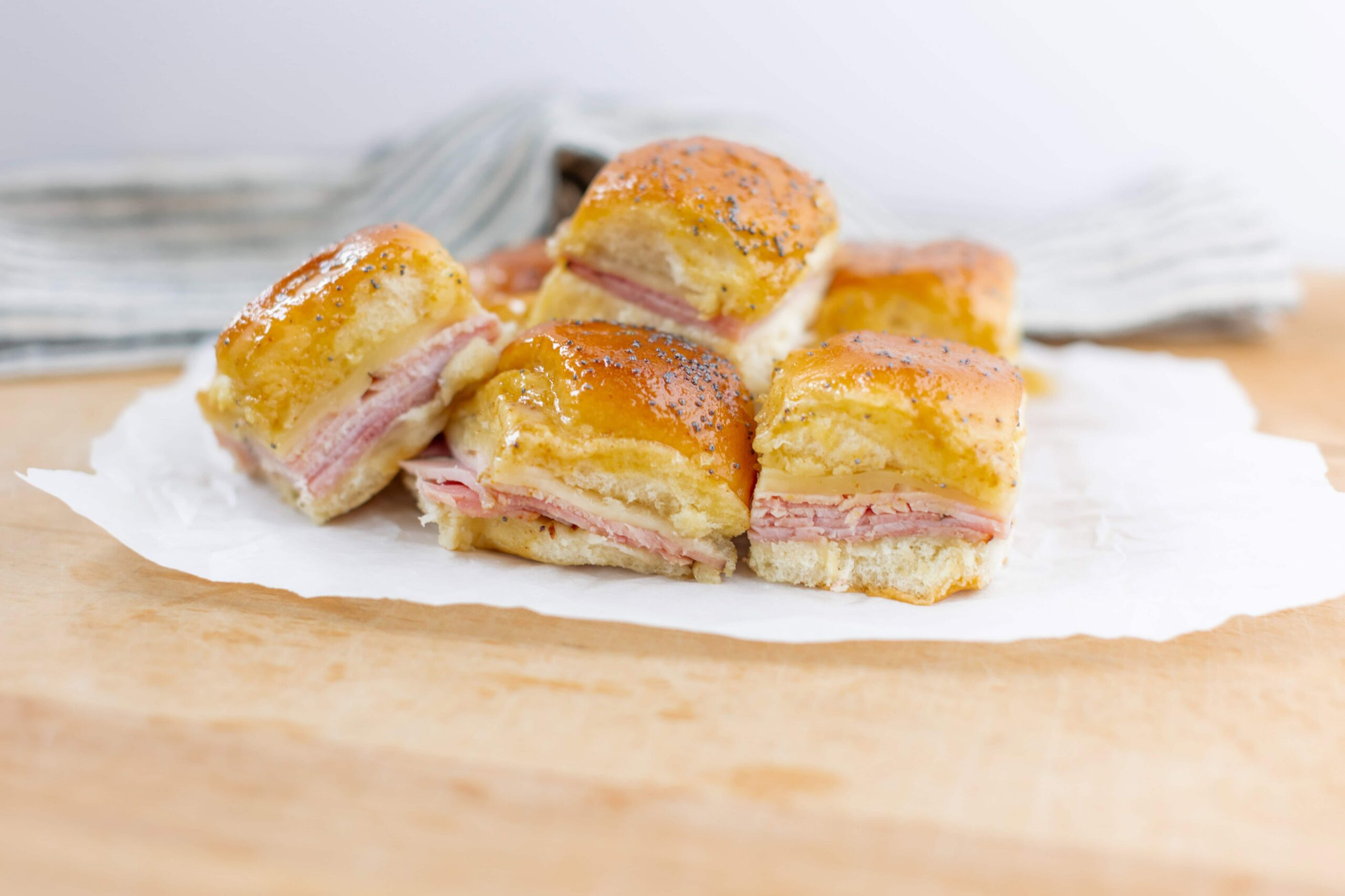 Buttered ham and cheese biscuits with a golden, flaky top, sprinkled with poppy seeds, served on parchment paper. Perfect for breakfast or snack, showcasing a savory pastry with ham and melted cheese filling.