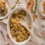 Creamy stuffed butternut squash casserole baked with herbs and topped with breadcrumbs, served in a white baking dish on a soft pink background with fresh rosemary sprigs.