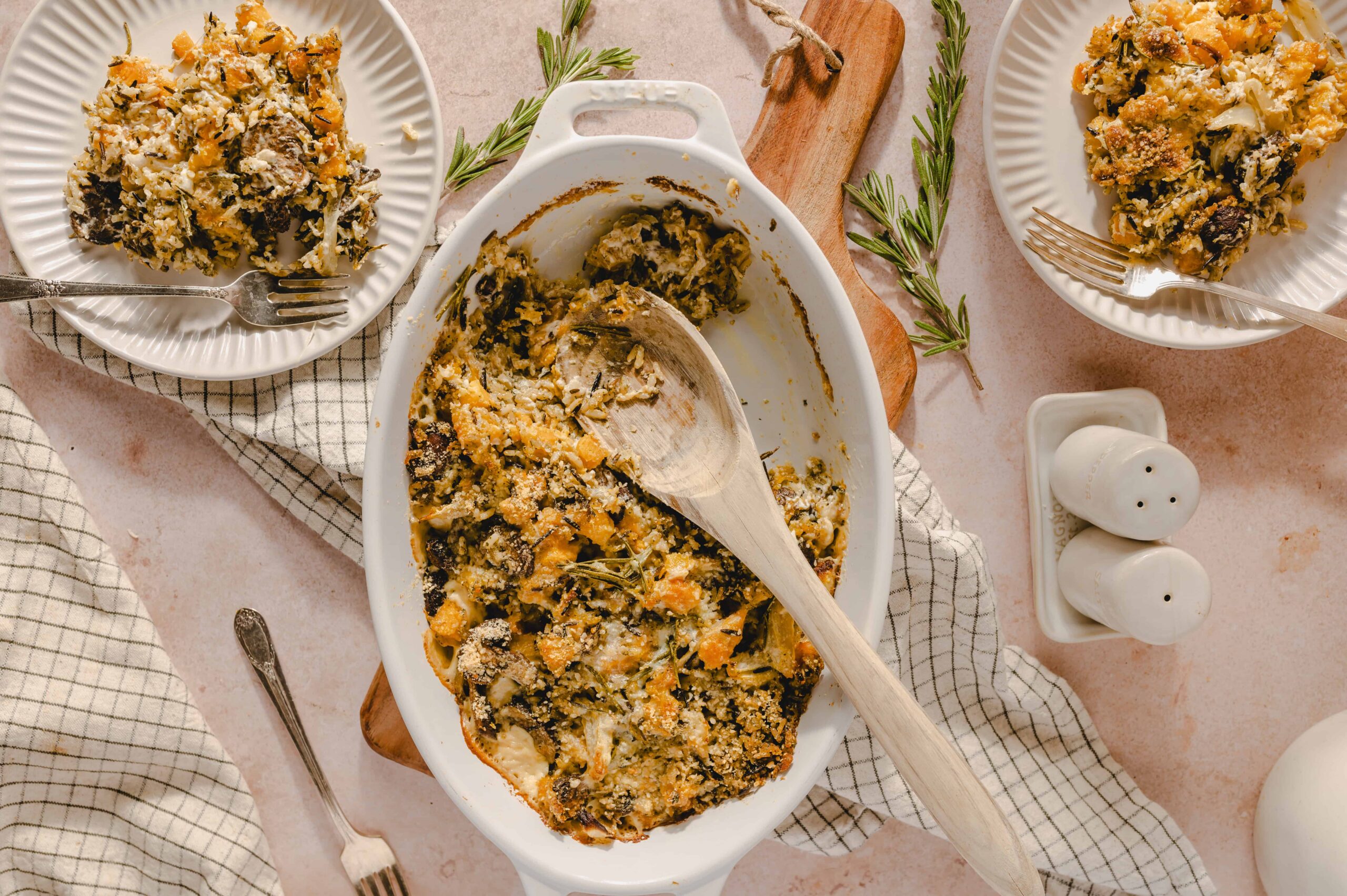 Creamy stuffed butternut squash casserole baked with herbs and topped with breadcrumbs, served in a white baking dish on a soft pink background with fresh rosemary sprigs.