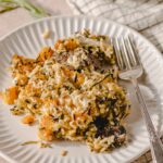 Creamy butternut squash and black rice casserole with herbs served on a white textured plate, garnished with herbs, placed on a beige surface with a fork and checkered napkin, perfect for warm comfort food.