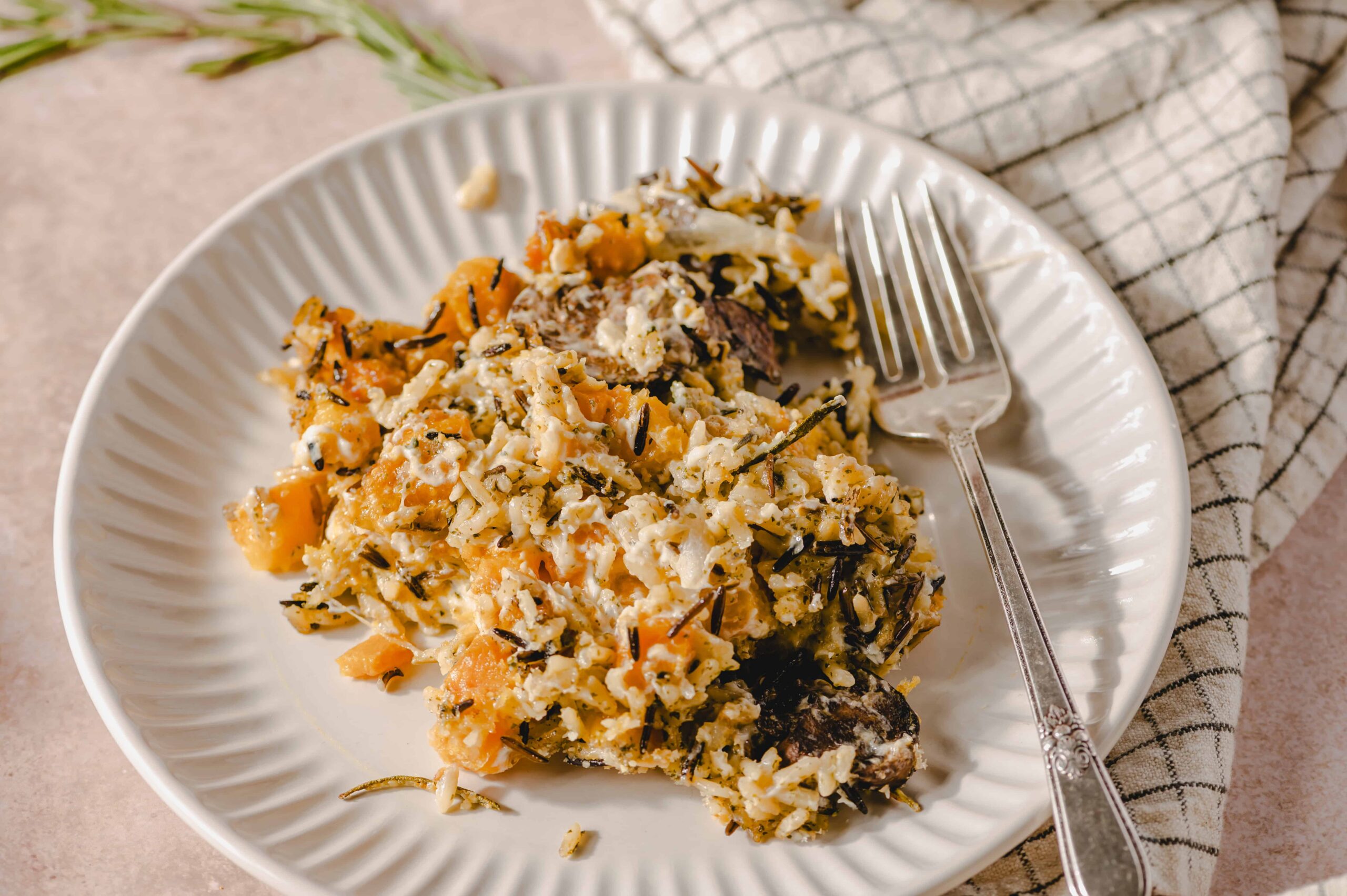 Creamy butternut squash and black rice casserole with herbs served on a white textured plate, garnished with herbs, placed on a beige surface with a fork and checkered napkin, perfect for warm comfort food.