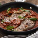 Succulent Italian veal marsala in tomato sauce garnished with basil leaves in a black skillet on wooden table, perfect for hearty Italian dinner.