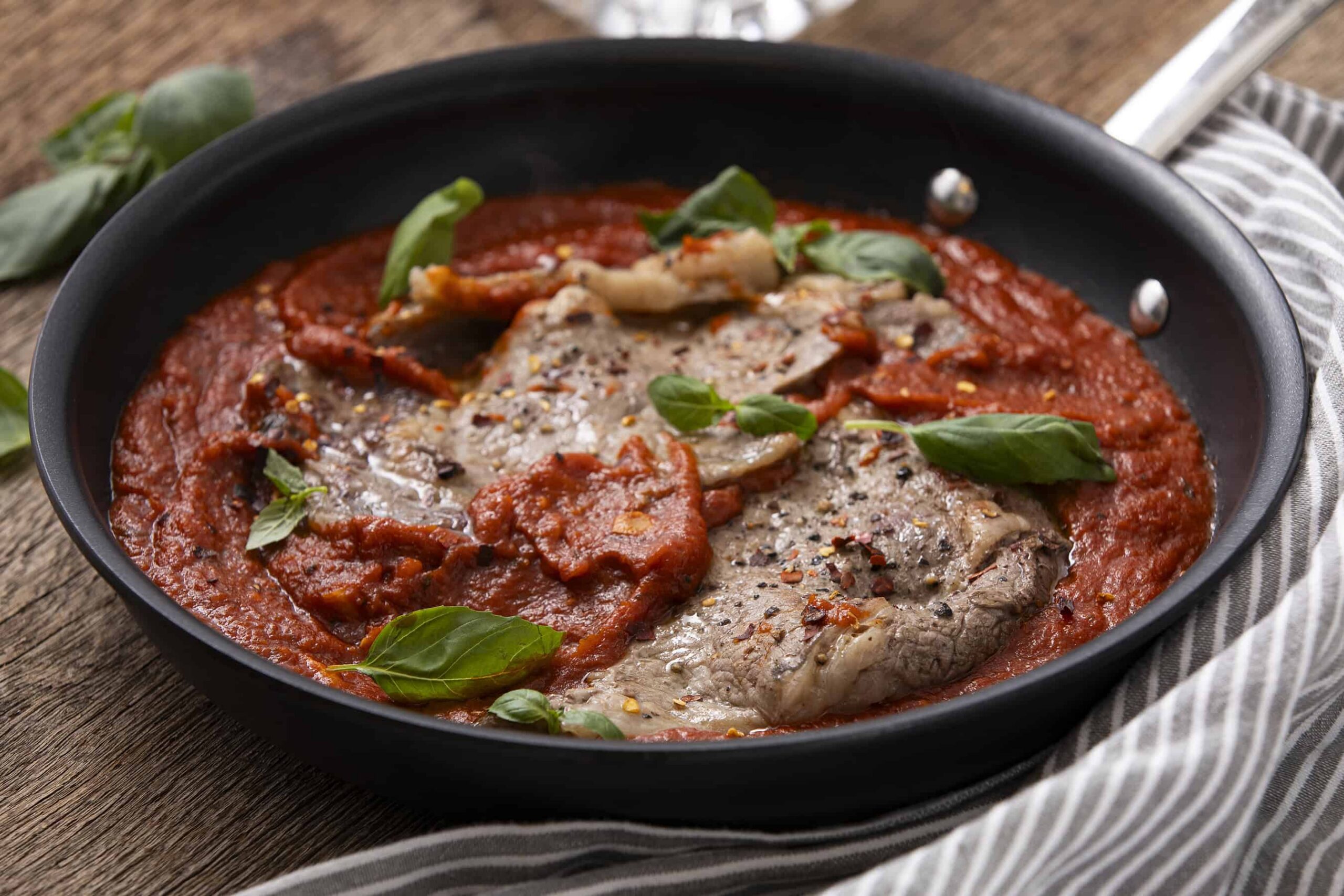 Succulent Italian veal marsala in tomato sauce garnished with basil leaves in a black skillet on wooden table, perfect for hearty Italian dinner.