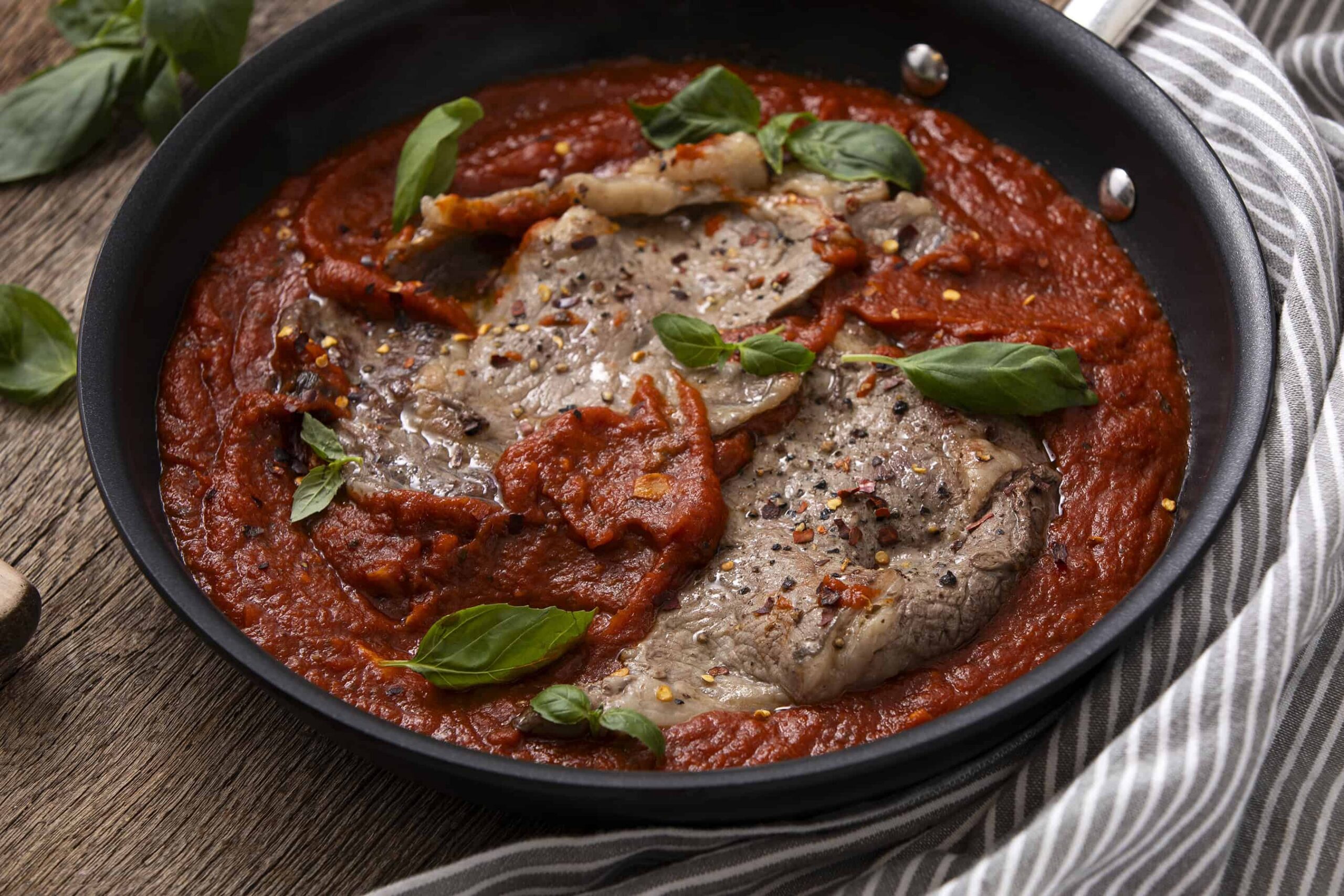 Succulent sliced beef in a rich tomato sauce garnished with fresh basil, served in a black skillet for an authentic Italian-style meat dish. Perfect for pasta or rice accompaniments.