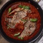 Juicy pork chops cooked in rich tomato sauce garnished with fresh basil and red pepper flakes, served in a black skillet for a flavorful and hearty meal.