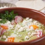 Sunny side up eggs in a rustic clay bowl with crumbled feta cheese, radish slices, fresh parsley, and crispy tortilla chips on the side, served as a delicious breakfast or brunch option.