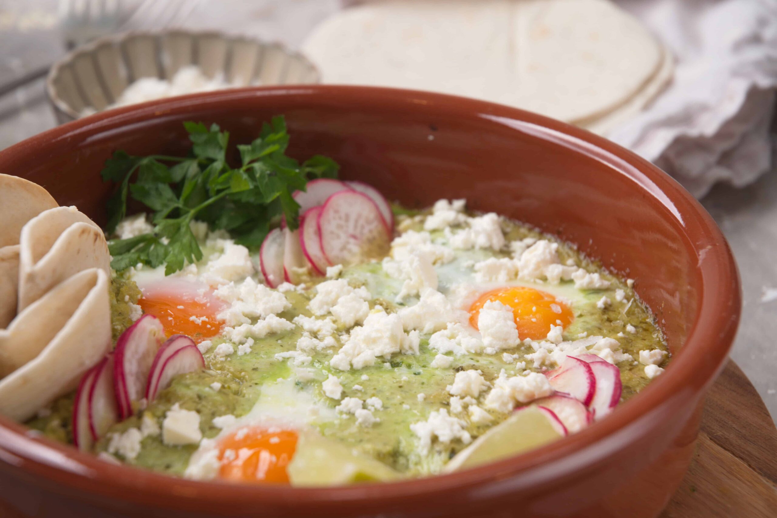 Sunny side up eggs in a rustic clay bowl with crumbled feta cheese, radish slices, fresh parsley, and crispy tortilla chips on the side, served as a delicious breakfast or brunch option.