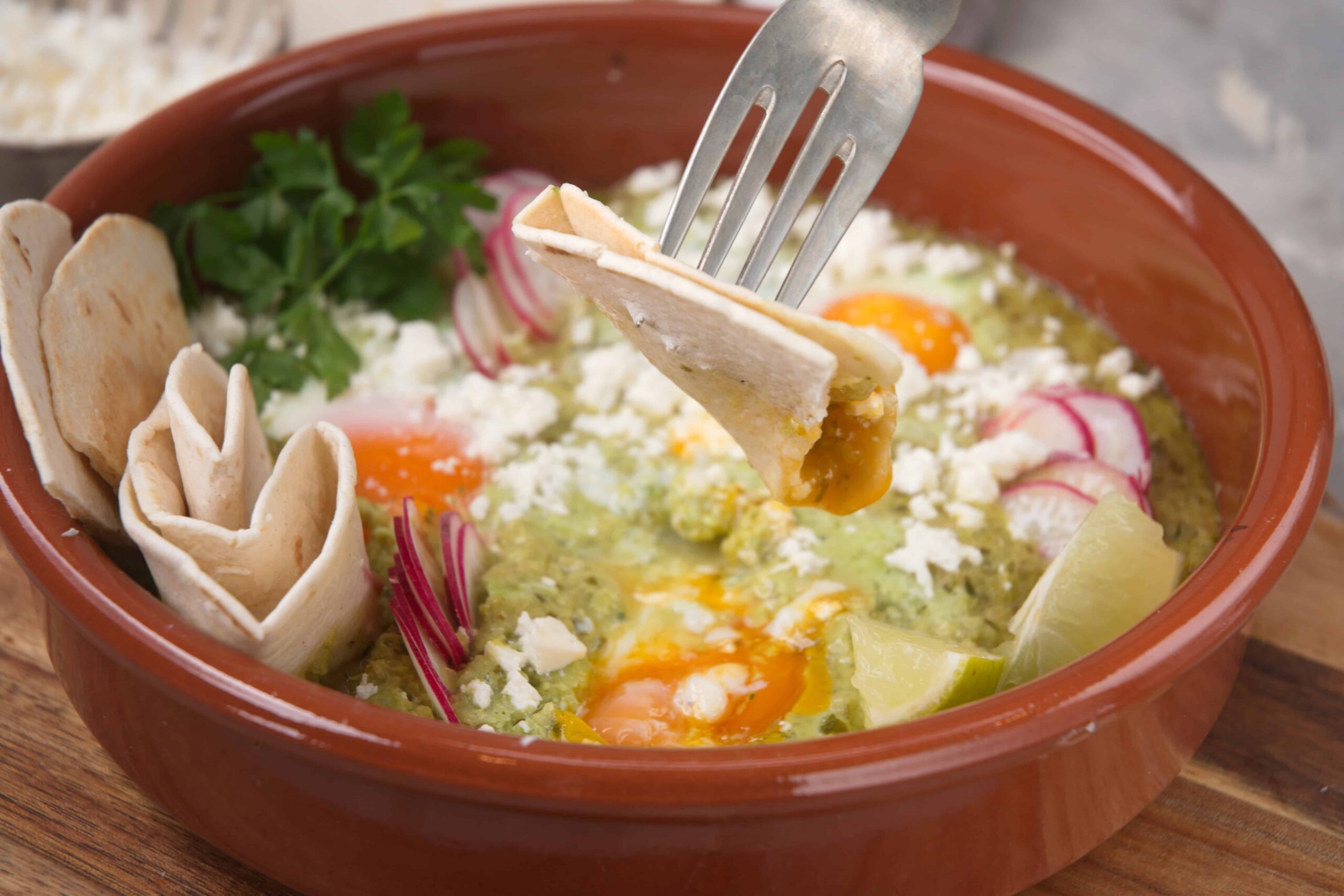 Fried eggs in a traditional Mexican dish served in a terracotta bowl with radishes, lemon, fresh herbs, and crispy tortillas, showcasing authentic Mexican breakfast comfort food.