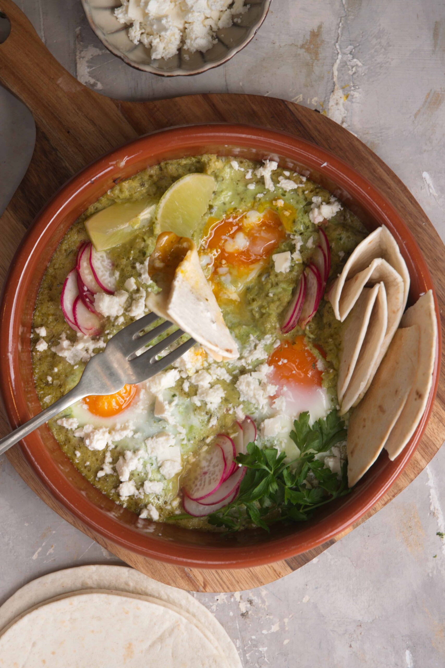 Creamy green chile breakfast casserole with baked eggs, sliced radishes, crumbled cheese, fresh cilantro, and warm flour tortillas on a wooden serving tray.