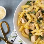 Creamy chicken pasta with spinach and parmesan cheese on a white plate, garnished with grated cheese, served with extra cheese in a small bowl and gold scissors on a blue surface.