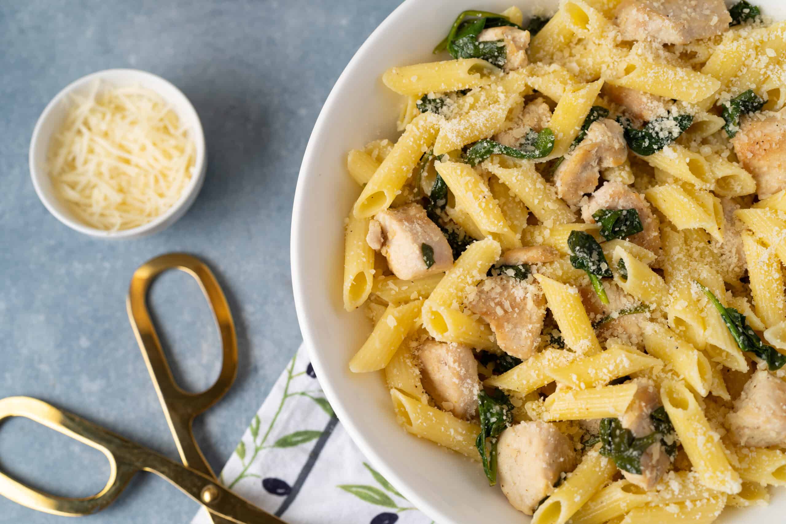 Creamy chicken pasta with spinach and parmesan cheese on a white plate, garnished with grated cheese, served with extra cheese in a small bowl and gold scissors on a blue surface.