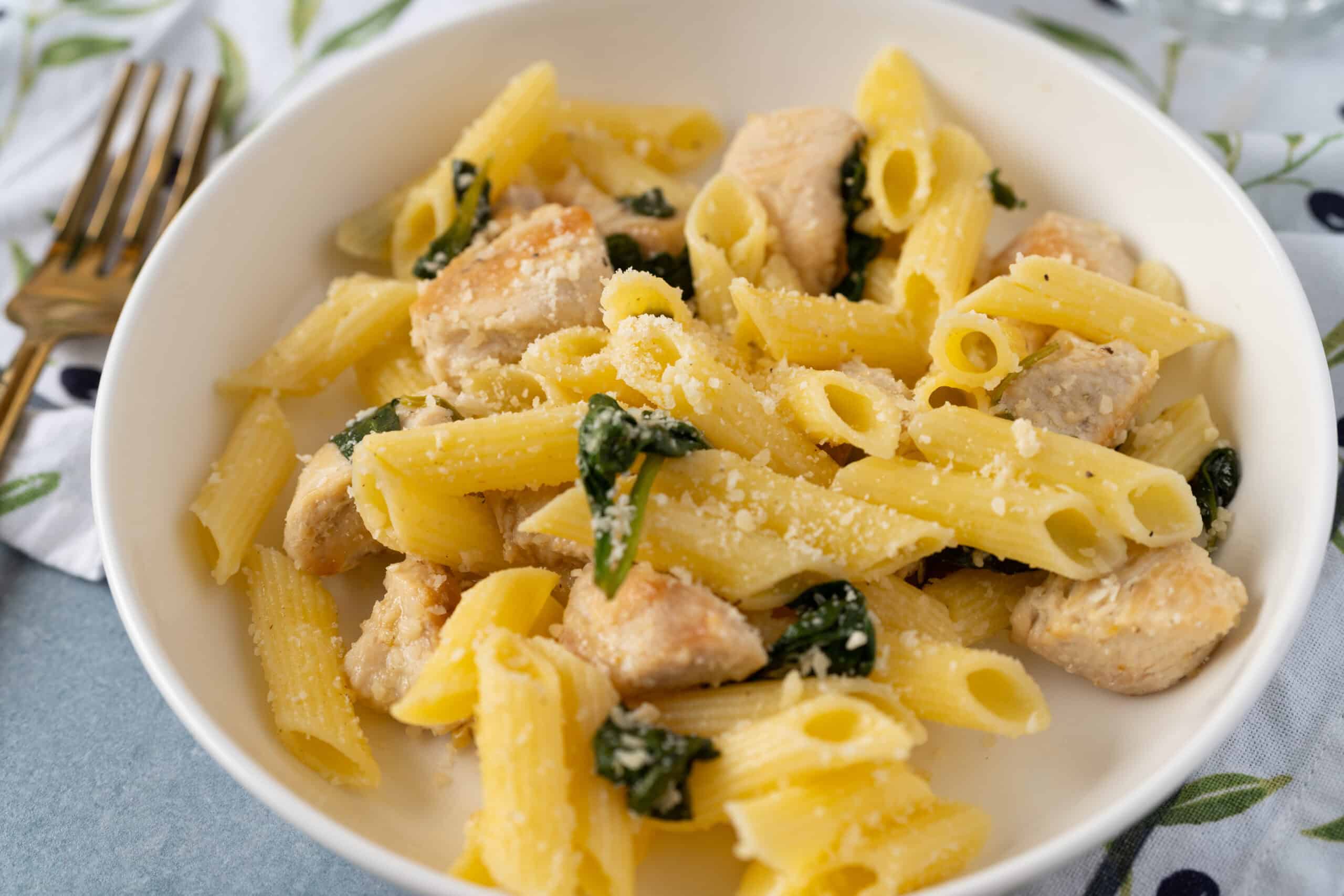 Creamy chicken pasta with spinach and grated Parmesan cheese on a white plate, served with a fork, fresh ingredients, and a colorful tablecloth. Perfect for quick weeknight dinners.