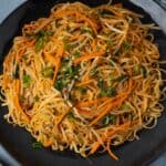 Stir-fried Asian noodle dish with vegetables, carrots, green onions, and bok choy served on a black plate. Perfect for an easy, flavorful lunch or dinner option, packed with fresh ingredients and savory flavors.