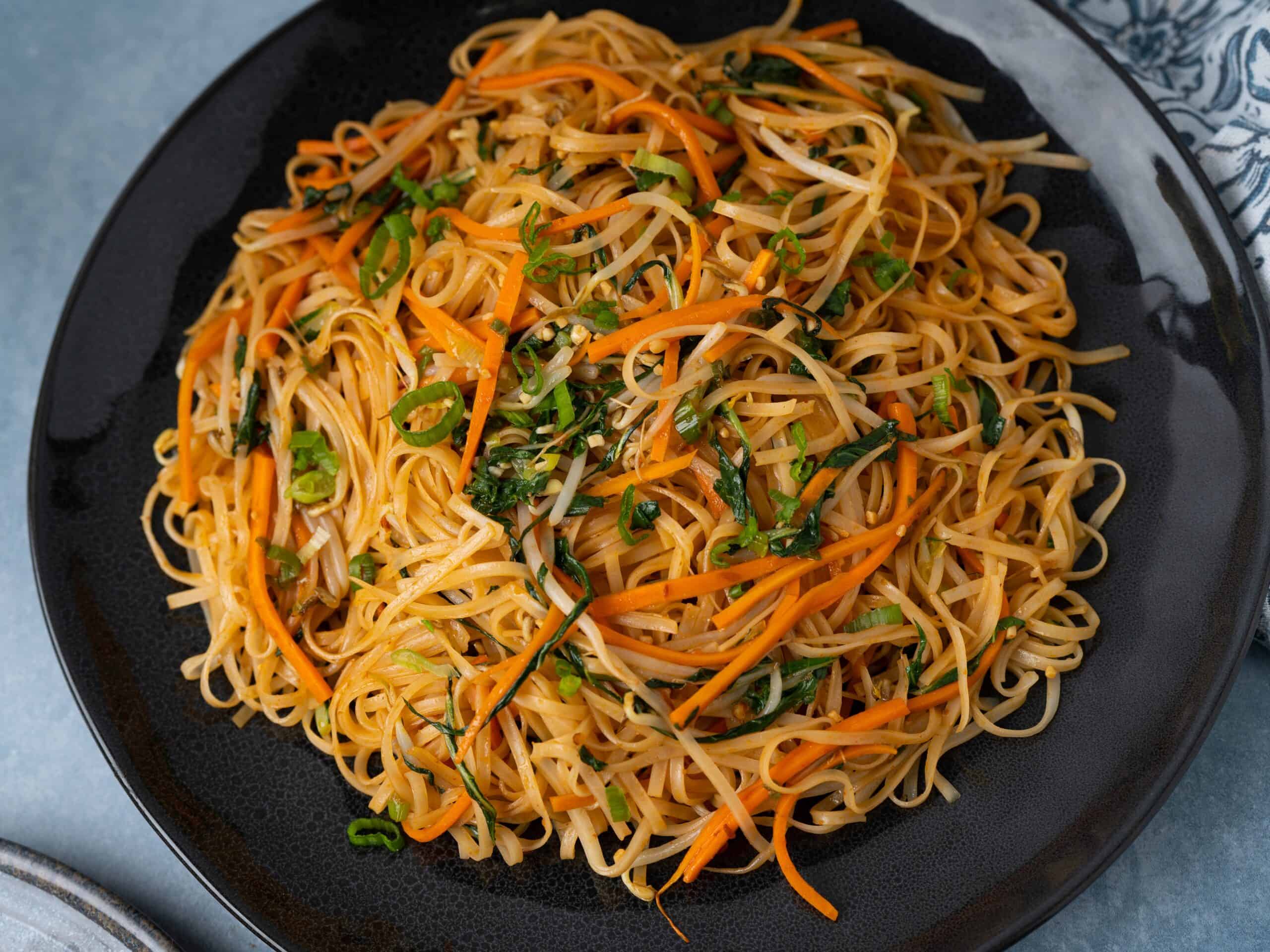 Stir-fried Asian noodle dish with vegetables, carrots, green onions, and bok choy served on a black plate. Perfect for an easy, flavorful lunch or dinner option, packed with fresh ingredients and savory flavors.