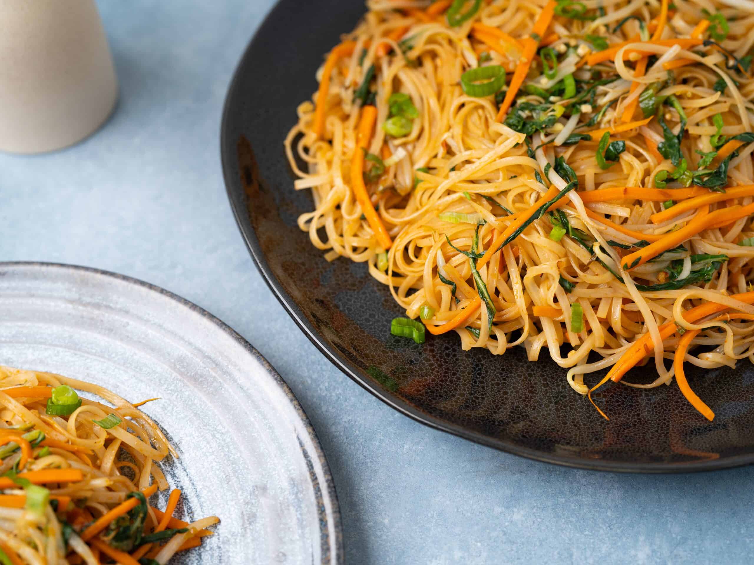 Stir-fried vegetable noodle dish with carrots, green onions, and bean sprouts on black and white plates, showcasing healthy vegetarian Asian cuisine.