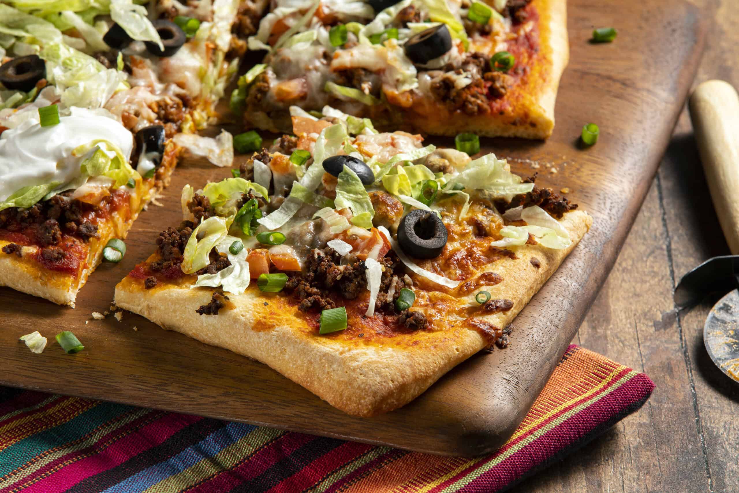 Freshly baked Mexican pizza with ground beef, lettuce, black olives, and cheese on a wooden serving board.